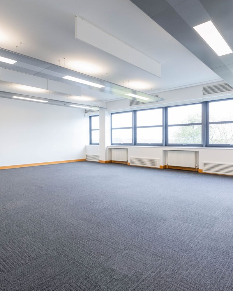 Large empty office space, with grey floor, a wall of large windows, a plain white wall, and lighting. 2 radiators to the far side. At HWIC.